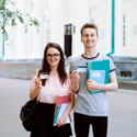 Two Best Friends Smiling, Holding Credit Cards And Showing Them To The Camera In Front Of Old University. Happy Students Use Advantages Of Electronic Cards In Everyday Life: Buy Goods, Pay Bills