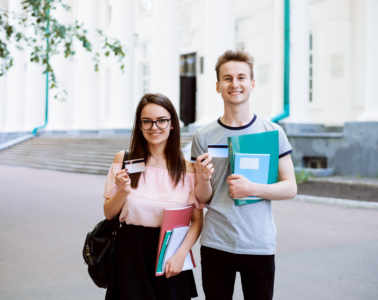 Two Best Friends Smiling, Holding Credit Cards And Showing Them To The Camera In Front Of Old University. Happy Students Use Advantages Of Electronic Cards In Everyday Life: Buy Goods, Pay Bills