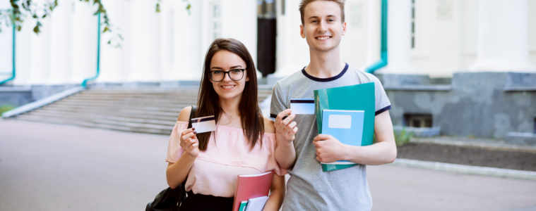 Two Best Friends Smiling, Holding Credit Cards And Showing Them To The Camera In Front Of Old University. Happy Students Use Advantages Of Electronic Cards In Everyday Life: Buy Goods, Pay Bills